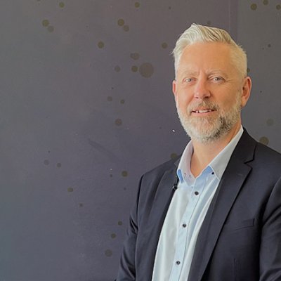 a man with a beard wearing a suit jacket stands in front of a grey wall